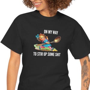 Könnte beinhalten: Schwarzes T-Shirt mit einer Cartoon-Illustration einer Frau, die einen Löffel hält, und dem Text "ON MY WAY TO STIR UP SOME SHIT" in Weiß. Die Frau trägt ein blaues Kleid und eine gelbe Schürze.