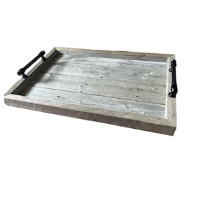 Puede incluir: Una bandeja rectangular de madera con un aspecto rústico y envejecido. La bandeja está construida con tablas de madera de color gris claro y presenta un asa negra mate oscura en cada extremo. La bandeja está diseñada para servir alimentos o bebidas.