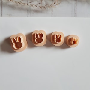Puede incluir: Cuatro cortadores de galletas beige con forma de conejito de diferentes tamaños. Cada cortador presenta un contorno de cara de conejito. Los cortadores están dispuestos en fila, el más grande a la izquierda y el más pequeño a la derecha. El fondo es de color claro y neutro.