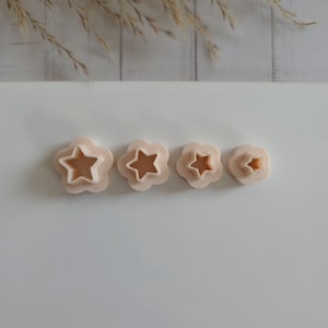 May include: Four star-shaped cookie cutters in varying sizes. Each cutter has a star shape within a flower shape, with a light beige color. The cutters are arranged in a row on a white surface, with dried plants in the background.