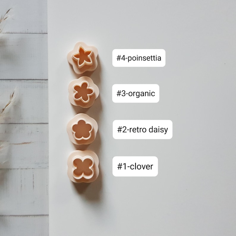 Pu&ograve; includere: Quattro formine per biscotti beige a forma di fiore. Le formine sono etichettate con i seguenti nomi: #4-poinsettia, #3-organic, #2-retro daisy e #1-clover. Le formine sono disposte verticalmente su una superficie bianca.