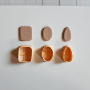 May include: Three light peach-colored clay shapes in rectangular, oval, and teardrop forms are arranged in a row above three matching light peach-colored plastic cutters. Each cutter features a cross-shaped internal support structure.