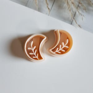 Peut inclure: Deux emporte-pièces en forme de croissant de lune beiges avec un motif de feuilles. Les emporte-pièces sont en plastique et sont placés sur une surface blanche. Le motif de feuilles est de couleur beige clair.