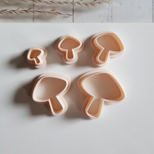 Puede incluir: Cinco cortadores de galletas con forma de hongo beige de varios tamaños están dispuestos sobre una superficie blanca. Los cortadores tienen un diseño simple y redondeado, perfecto para hornear o hacer manualidades. El fondo incluye algunos tallos secos.