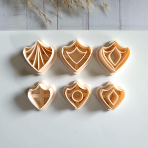 Könnte beinhalten: Sechs beige Ausstecher aus Kunststoff in verschiedenen Schildformen. Die Ausstecher haben detaillierte Designs, darunter geometrische Muster und ein Kreuz. Die Ausstecher sind auf einer weißen Oberfläche angeordnet.