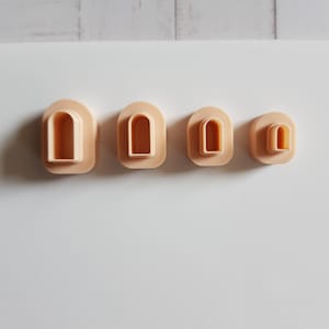 Op de afbeelding: Vier beige polymeerklei uitsteekvormen in een boogvorm, gerangschikt van groot naar klein. Elke uitsteekvorm heeft een rechthoekige basis met afgeronde hoeken en een holle booguitsparing. De uitsteekvormen zijn ontworpen voor knutselen.