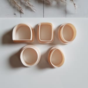 May include: Five beige-colored cookie cutters in various shapes: a rounded arch, a rectangle with rounded corners, a circle, a circle, and an oval. The cutters are arranged on a white surface, with dried plants in the background.