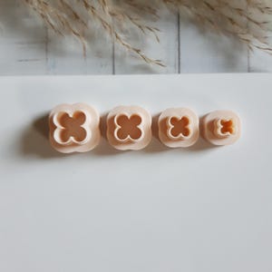 Può includere: Quattro formine per biscotti a forma di fiore color pesca chiaro di varie dimensioni. Le formine sono disposte in fila, con la più grande a sinistra e la più piccola a destra. Le formine hanno un design arrotondato a quattro petali.