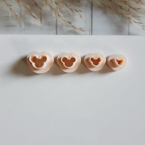Peut inclure: Quatre emporte-pièces en forme de cœur de différentes tailles, chacun avec un motif de tête de Mickey Mouse au centre. Les emporte-pièces sont de couleur pêche clair et sont disposés en rangée sur une surface blanche.