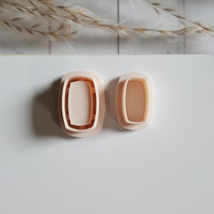 May include: Two light peach-coloured rectangular cookie cutters. The cutters have a rounded rectangular shape with a recessed interior. The larger cutter is on the left, and the smaller cutter is on the right. The background is white.