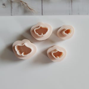 Puede incluir: Cuatro cortadores de galletas en forma de pétalo de color rosa claro con un diseño de hoja de ginkgo. Los cortadores están dispuestos sobre una superficie blanca. La forma de la hoja de ginkgo es visible en el centro de cada cortador. Los cortadores son de plástico.