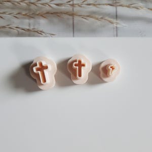 Può includere: Tre formine per biscotti a forma di croce rosa chiaro di diverse dimensioni. Le croci sono in rilievo all'interno di una forma arrotondata, simile a una nuvola. Lo sfondo è bianco tenue, con un accenno di fogliame secco in alto.