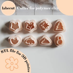 Op de afbeelding: Een verzameling lichtroze polymeerklei-uitsteekvormen in verschillende vormen, waaronder een vos, ster, boom, vogel en berg. De afbeelding bevat de tekst "labecut Cutter for polymer clay" en "STL file for 3D printer."