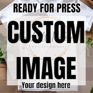 May include: White t-shirt with the text "READY FOR PRESS" at the top. Below, large black text reads "CUSTOM IMAGE" with "Your design here" at the bottom. A circular design with "DTF TRANSFERS" is in the center.