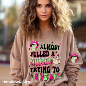 Op de afbeelding: Een beige sweatshirt met de tekst "ALMOST PULLED A MUSCLE TRYING TO GIVE AF" in zwart, roze en groen. Het ontwerp bevat een middelvinger, een geblokte bliksemschicht en cartoonlippen met een geblokte tong.