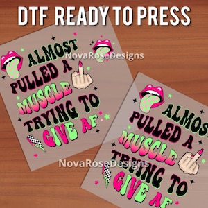 Op de afbeelding: Twee DTF (Direct to Film) ready-to-press transfers met de tekst "ALMOST PULLED A MUSCLE TRYING TO GIVE AF" in zwart, neonroze en groen. Het ontwerp bevat een middelvinger, lippen en sterren.