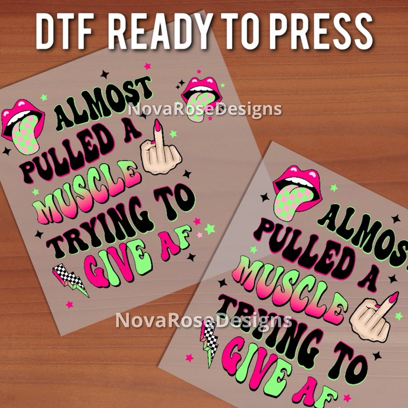 Op de afbeelding: Twee DTF (Direct to Film) ready-to-press transfers met de tekst "ALMOST PULLED A MUSCLE TRYING TO GIVE AF" in zwart, neonroze en groen. Het ontwerp bevat een middelvinger, lippen en sterren.