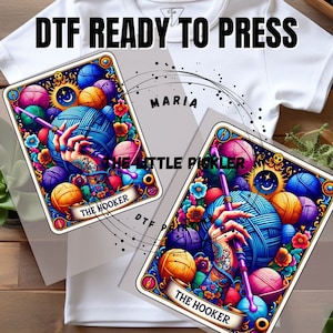 May include: Two colorful tarot cards with the text "The Hooker" and "Maria The Little Pickler" on them. The cards feature a hand holding a knitting needle and surrounded by yarn balls.