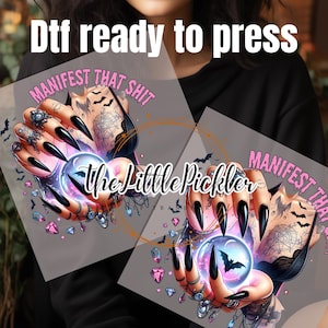 May include: Two digital transfers with a gothic aesthetic. Both feature a hand with long black nails holding a crystal ball with a bat inside. The text "MANIFEST THAT SHIT" is on the top of each transfer.