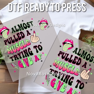 Op de afbeelding: DTF ready to press transfer met de tekst "ALMOST PULLED A MUSCLE TRYING TO GIVE AF" in een retro lettertype. Het ontwerp bevat een middelvinger, lippen met een geblokte tong, sterren en een bliksemschicht. Het ontwerp is in neonroze, groen en zwart.