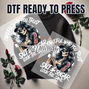 May include: A digital transfer design featuring a skeleton woman with long black hair, wearing a black hat and sunglasses, holding a bottle. The design includes the text "Careful who you trust, Salt & Sugar look the same".