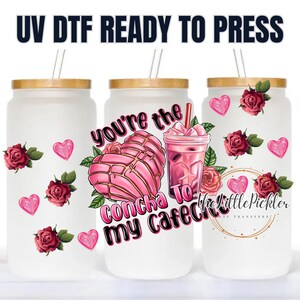 May include: A white tumbler with a wooden lid and a straw. The tumbler has a pink and white illustration of a concha bread, a glass of cafe con leche, and the text "You're the Concha to my Cafe con Leche".