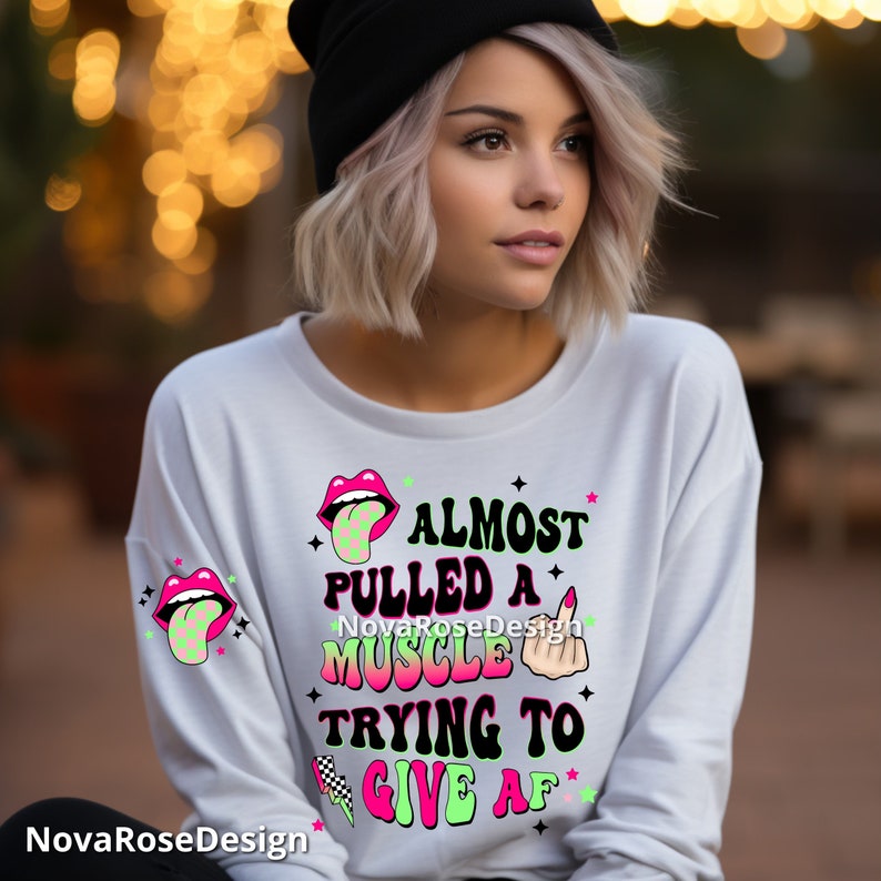 Op de afbeelding: Witte longsleeve met de tekst "ALMOST PULLED A MUSCLE TRYING TO GIVE AF" in zwart, roze en groen. Het shirt heeft grafische ontwerpen van lippen, een middelvinger en een geblokte vlag.
