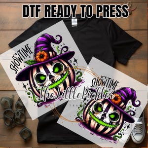 May include: DTF Ready to Press design featuring a Halloween-themed pumpkin with a witch hat and the text "It's Showtime". The pumpkin has green eyes and a green grin. The design is on a transparent sheet, ready for application.