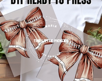 May include: Two brown football-themed bows with white stitching and white stripes. The bows are on a white t-shirt with the text "DTF READY TO PRESS" on the top. The text "THE LITTLE PICKLE" is visible in the background.