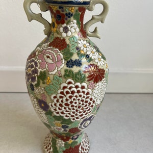May include: A white ceramic vase with a floral design. The vase has two handles and a wide base. The flowers are in shades of pink, red, green, blue, and white.