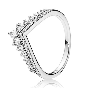 May include: A silver ring with a V-shaped design. The ring is adorned with small, clear gemstones along the V-shape and larger gemstones at the top. The band is smooth and polished, reflecting light.
