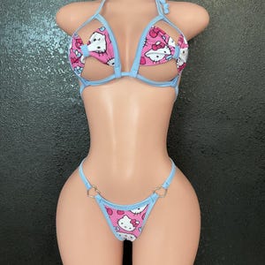 May include: A two-piece bikini set featuring a light blue trim and a pink fabric patterned with a cartoon cat design. The top has a halter neck and open bust design. The bottom has heart-shaped ring accents.