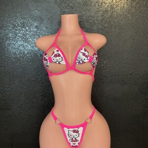 May include: A pink and white bikini set featuring a Hello Kitty print. The halter top and thong-style bottoms have pink trim and heart-shaped metal accents. The bikini is displayed on a mannequin.