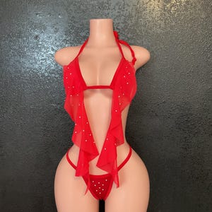 May include: Red lingerie set with a halter top, a flowing fabric panel, and a thong-style bottom. The set is adorned with small, white embellishments. The lingerie is displayed on a mannequin against a dark background.