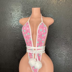 May include: Pink bunny-themed bodysuit with a halter neck and white trim. The fabric features a pattern of snowflakes and candy canes. The waist is cinched with a white tie, and two white pom-poms are attached at the bottom.