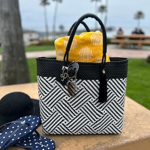 May include: Black and white woven tote bag with a yellow and white patterned towel inside. The bag has a black tassel and a gold charm with the letter "P" on it. Sunglasses are also inside the bag.