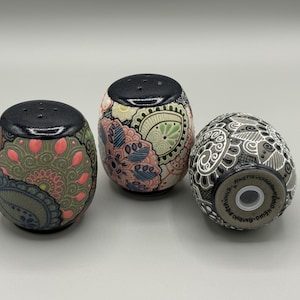 May include: Three decorative ceramic salt and pepper shakers. The shakers are egg-shaped with black tops and intricate floral and paisley designs in shades of pink, green, blue, and grey. The shakers are on a light grey surface.