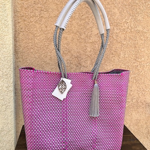 Puede incluir: Un bolso tote grande de color rosa y plateado con un borla gris y asas de cuero blanco. El bolso está hecho de plástico tejido y tiene un amuleto en forma de hoja.