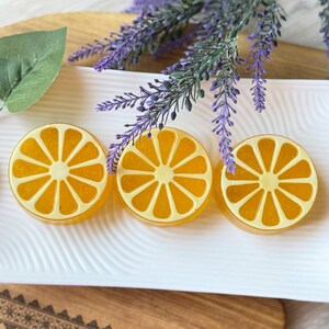 Può includere: Tre saponi rotondi e traslucidi a forma di fetta di agrume arancione, con scorza e segmenti giallo pallido, disposti in fila su un vassoio rettangolare bianco dalla consistenza ondulata, accompagnati da rametti di lavanda viola.
