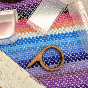 May include: A green and purple succulent-shaped soap and a smaller succulent soap on a wooden slice. Clear plastic containers and a wooden ring are on a colourful beaded surface. The background is a white-framed wooden surface.