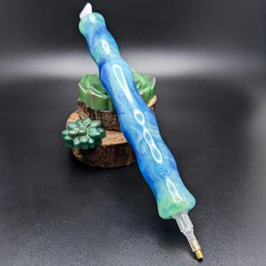 May include: A diamond painting stylus featuring a marbled blue and green resin handle with a wavy, ergonomic shape. The tool includes a clear plastic tip attachment with a small metal point at the end, resting against a dark green succulent base.