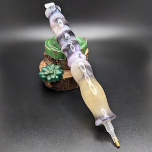 May include: A diamond painting pen featuring a contoured, multi-sectioned handle with a swirling pattern of translucent purple, white, and gold glitter resin. The tool has a clear plastic tip attachment at the bottom and a metallic gold point.