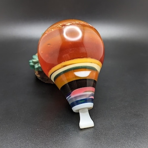 May include: A teardrop-shaped glass ornament featuring a smooth, glossy finish with a vibrant colour palette. The top section is a deep amber orange, transitioning into horizontal bands of yellow, green, black, pink, and blue, ending in a white tip.