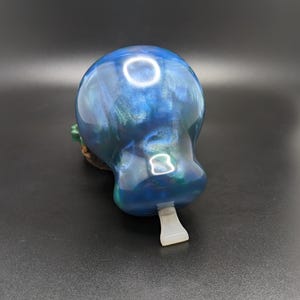 May include: A smooth, rounded object with a glossy finish in shades of deep blue, teal, and iridescent green. The surface features swirling, pearlescent patterns and a small, white, rectangular tab protruding from the bottom edge of the piece.