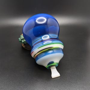 May include: A rounded glass object with a smooth, deep blue top section that transitions into a base featuring horizontal bands of white, light blue, green, and thin red stripes. A small, white, flat, rectangular tab extends from the bottom.