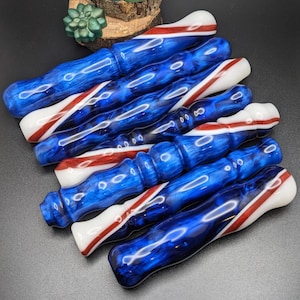 May include: A selection of resin pen blanks in a patriotic design. The blanks are predominantly blue with white and red accents. The glossy finish and swirling patterns create a decorative effect, suitable for crafting pens.