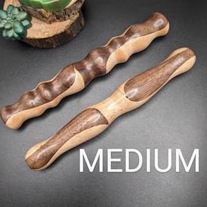 May include: Two wooden massage tools with a two-tone design. The tools are labelled "MEDIUM" in white text. One tool has a wavy handle, and the other has a smooth, curved handle. The wood has a light and dark brown colour.
