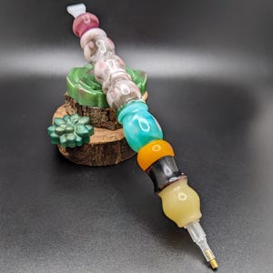 May include: A decorative diamond painting pen featuring a series of colourful beads in pink, clear, teal, orange, black, and yellow. The pen is displayed on a small wooden base with a green succulent and a green leaf-shaped object.