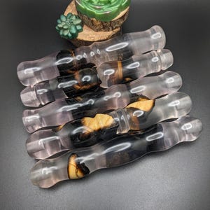 May include: Several clear, pink-hued resin wands with dark wood and amber inclusions are displayed. The wands are arranged diagonally against a dark backdrop. A small green succulent and a green soap are in the background.