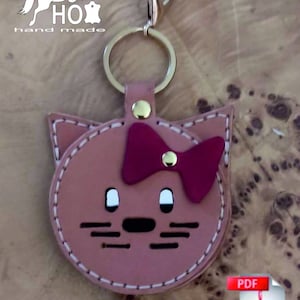 May include: A pink leather cat-shaped keychain with a red bow and white eyes. The keychain has a gold metal ring and a gold metal clasp. The text "PATTERN" is printed below the keychain. The text "PDF" is printed on a red and white icon of a document with a bent corner.